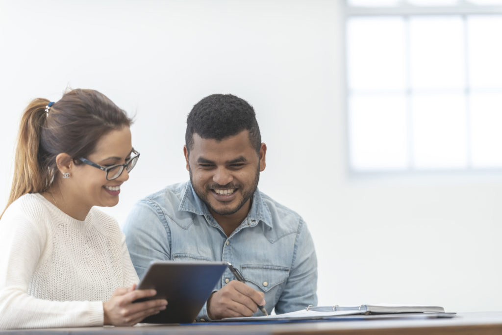 A couple are sitting at a table in their home and looking at an electronic tablet. There are papers on the table in front of them.